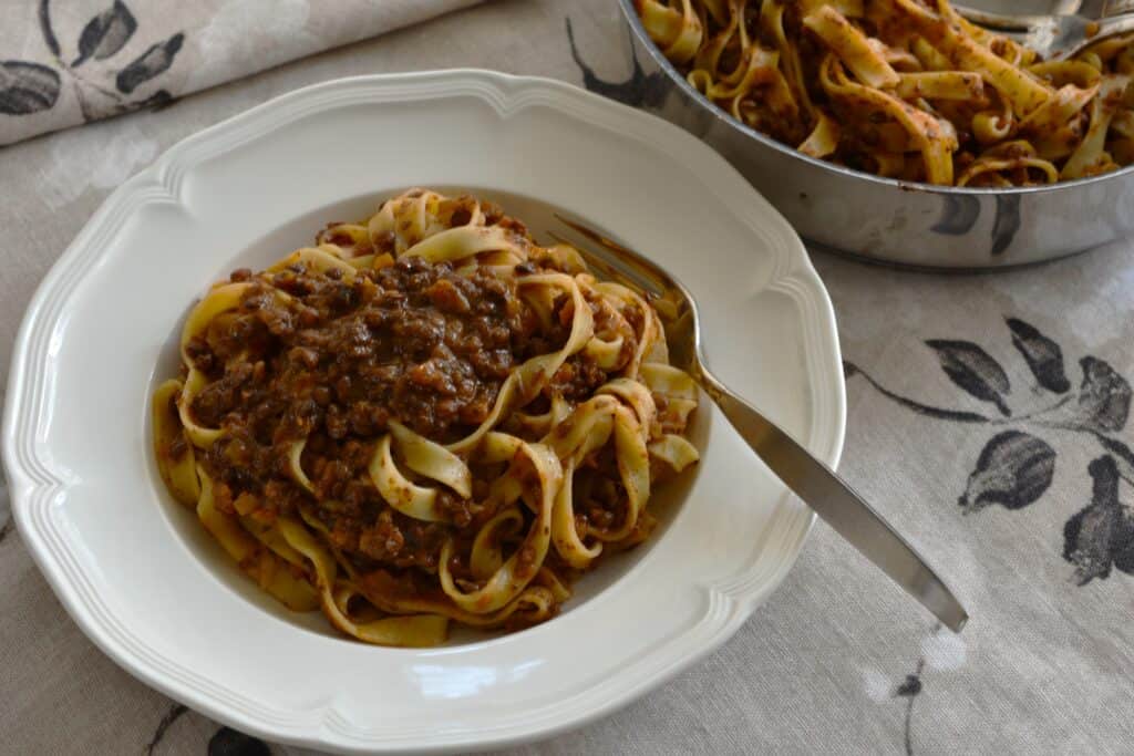 Vegan ragu with tagliatelle on a white plate with a fork and a pan in the background.