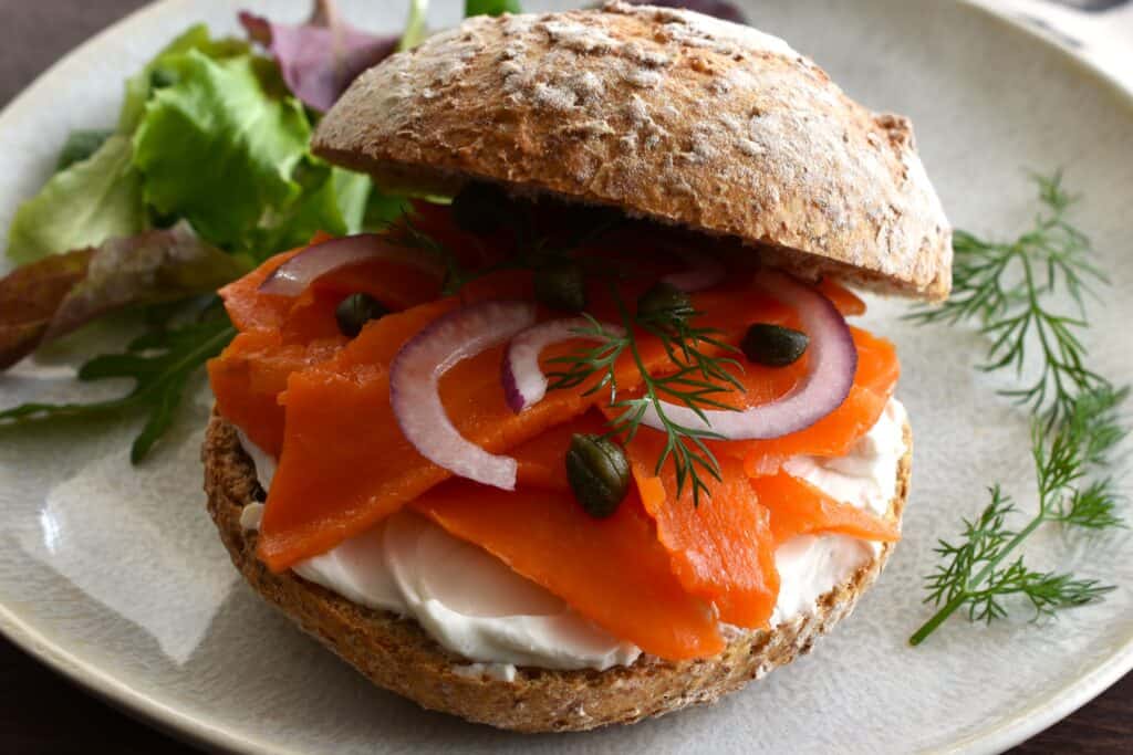 A close-up image of vegan smoked salmon and cream cheese in a bread roll on a plate with a garnish of dill and mixed leaf salad.