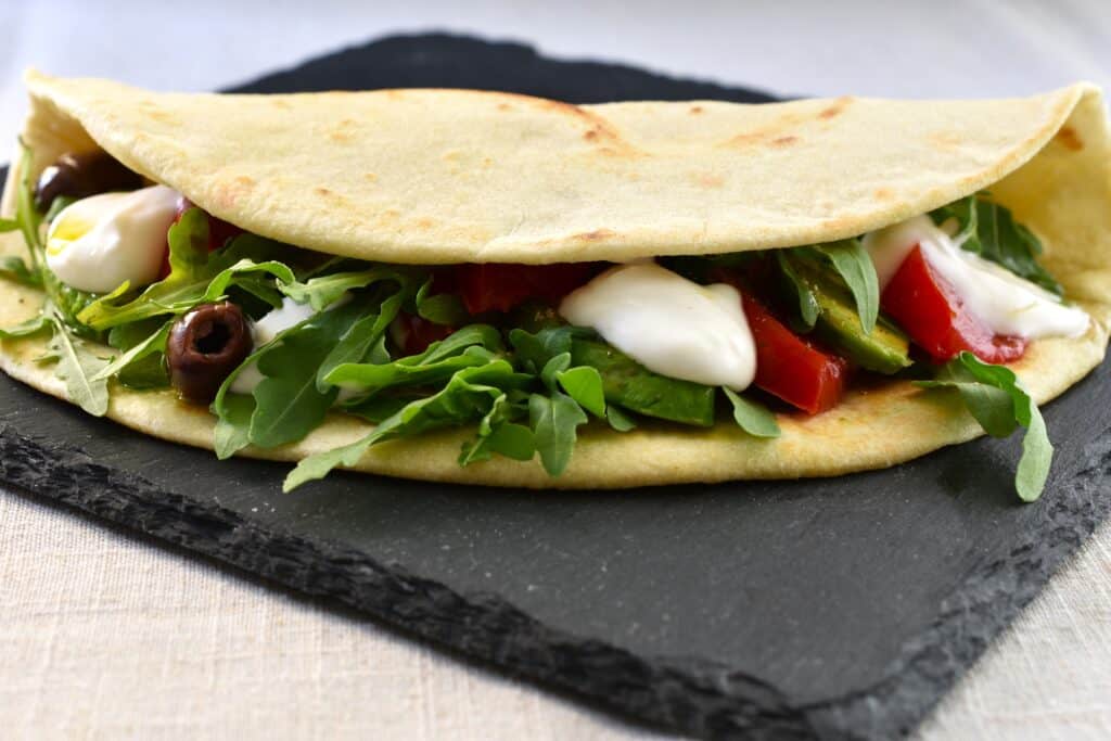 A vegan piadina folded in half, with a filling of avocado, tomato, vegan stracchino, rocket and olives.