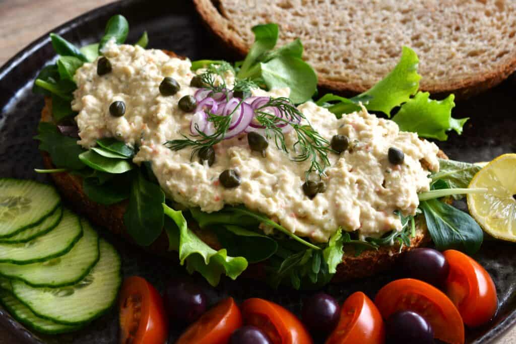 A vegan tuna paté sandwich with a garnish of red onion, dill, capers, cucumber, tomatoes and black olives.