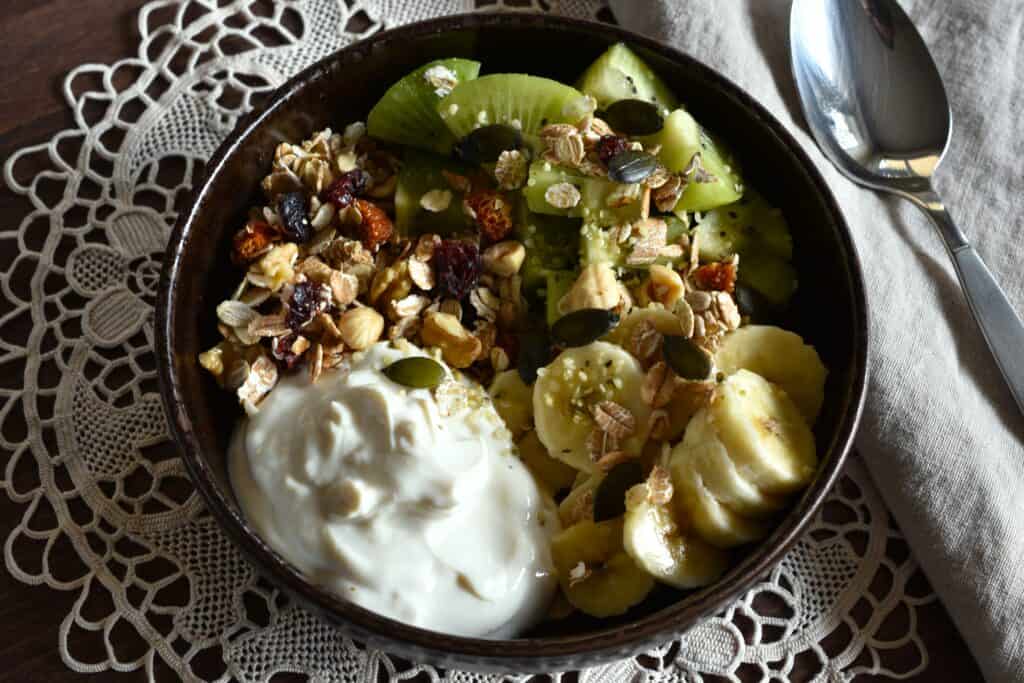 Raw vegan granola with kiwi, banana, soya yoghurt, pumpkin seeds, and hemp seeds in a brown bowl.