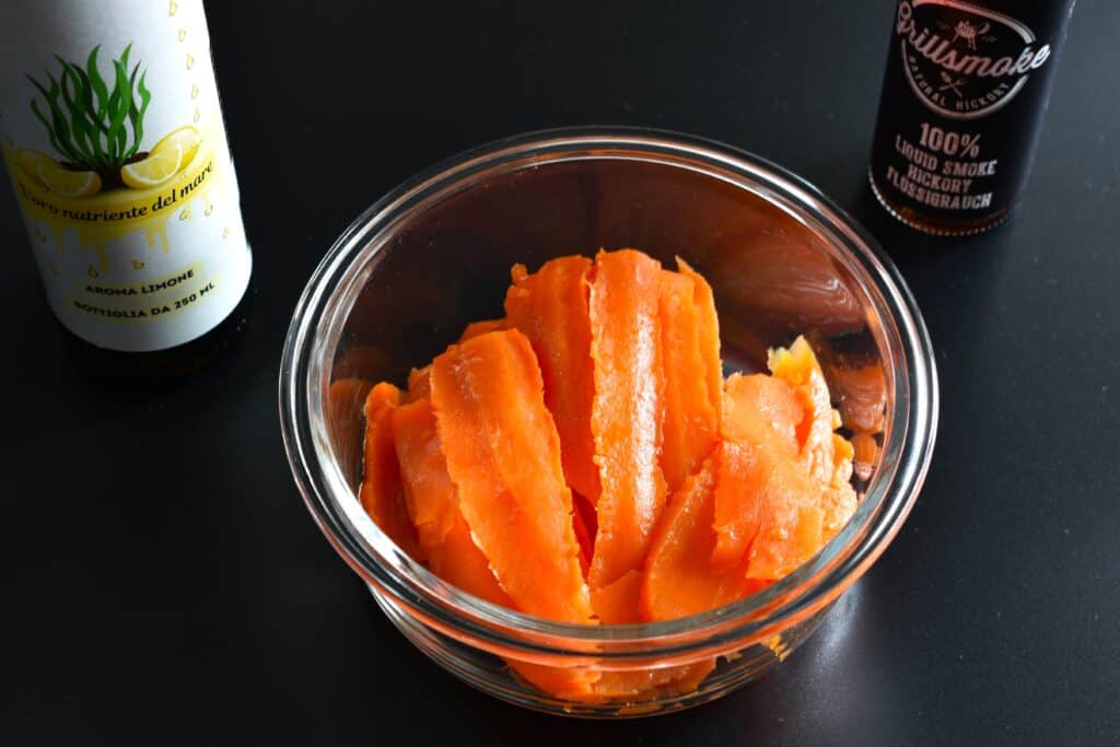 Slices of marinated carrot in a glass bowl with a bottle of Nutra Olisear and liquid grillsmoke.