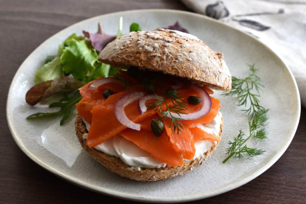 Vegan smoked salmon and vegan philadelphia in a bread roll on a plate with a garnish of dill and mixed leaf salad.