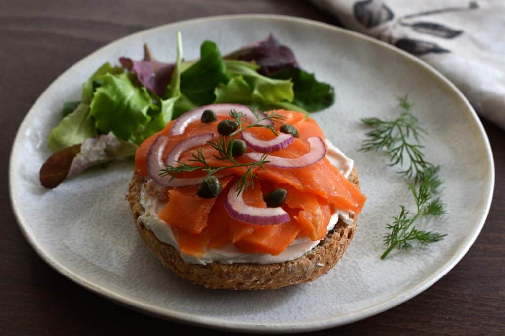 Vegan smoked salmon and vegan philadelphia with red onion and capers on the base of a bread roll on a plate with a garnish of dill and mixed leaf salad.