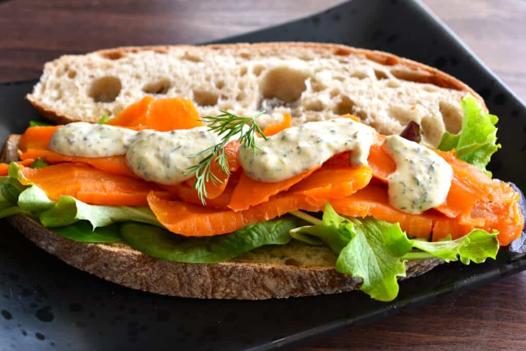 Vegan smoked salmon on sourdough bread with mixed leaf salad and vegan dill, "honey" and mustard sauce on a black plate.