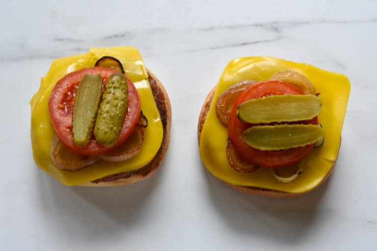 Vegan burger bun bases topped with vegan burgers, vegan cheese, fried onion, a slice of tomato, and slices of gherkin.