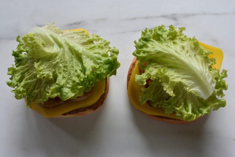 Vegan burger bun bases topped with vegan burgers, vegan cheese, fried onion, a slice of tomato, slices of gherkin and lettuce leaves.