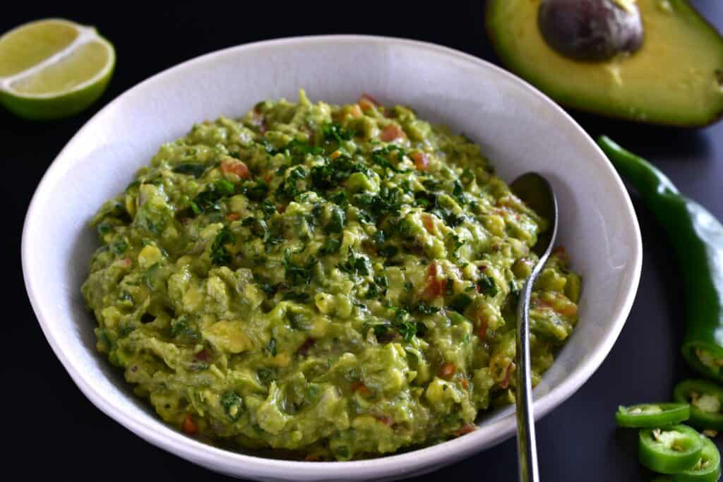 Vegan guacamole in a white bowl with a spoon and recipe ingredient props.