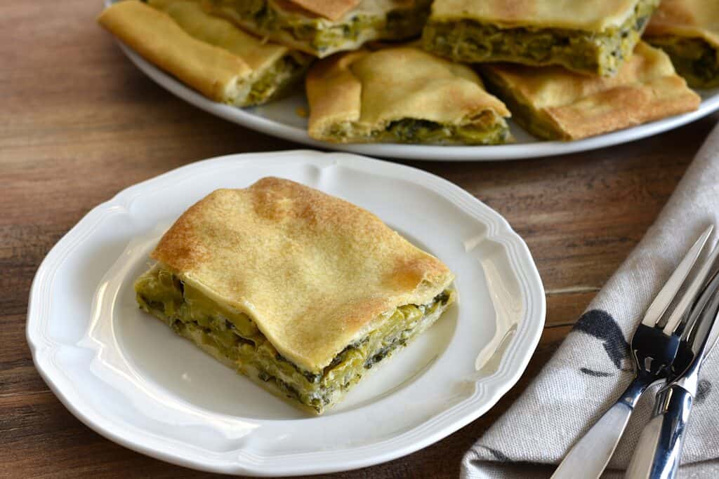 A piece of Ligurian torta verde on a white plate with other pieces of torta verde on a serving plate in the background.