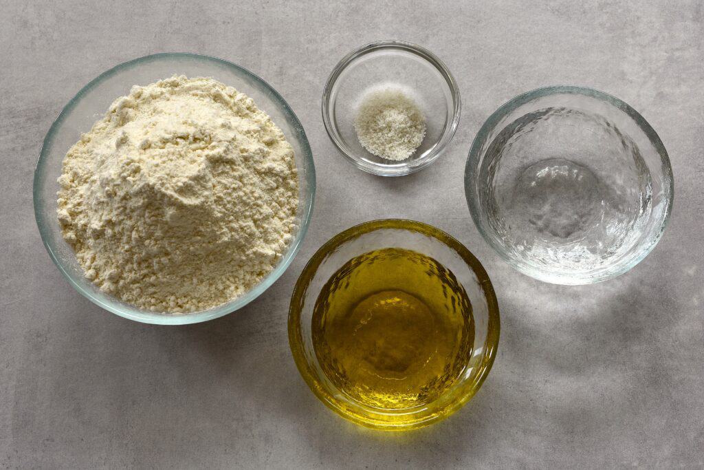 Flour, water, olive oil, and salt (ingredients for pasta matta) in individual glass bowls on a worktop.