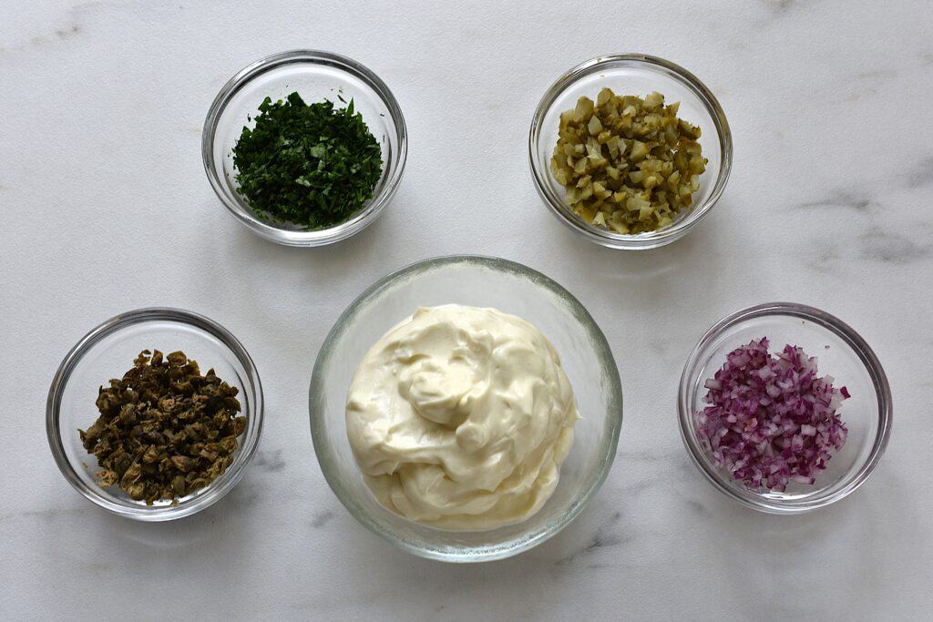 Ingredients for tartare sauce: Vegan mayonnaise, chopped onion, capers, gherkins, and parsley in glass bowls.