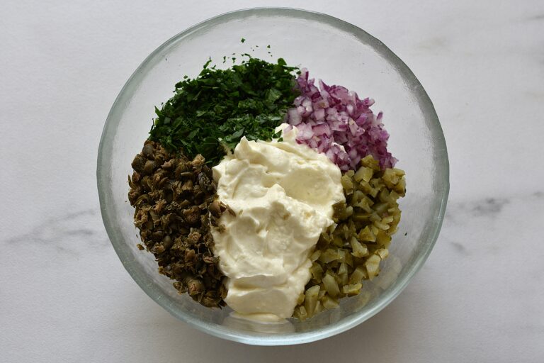 Vegan mayonnaise, chopped onion, capers, gherkins and parsley (ingredients for vegan tartare sauce) in a glass bowl.