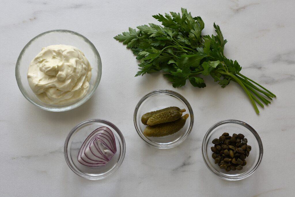 Vegan mayonnaise, onion, capers, gherkins, and parsley on a worktop.