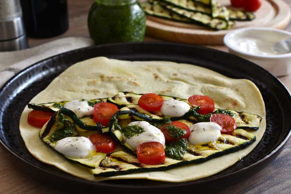 A vegan piadina with a filling of grilled courgettes (zucchini), tomatoes, vegan stracchino cheese, vegan basil pesto. olive oil, salt, and black pepper.