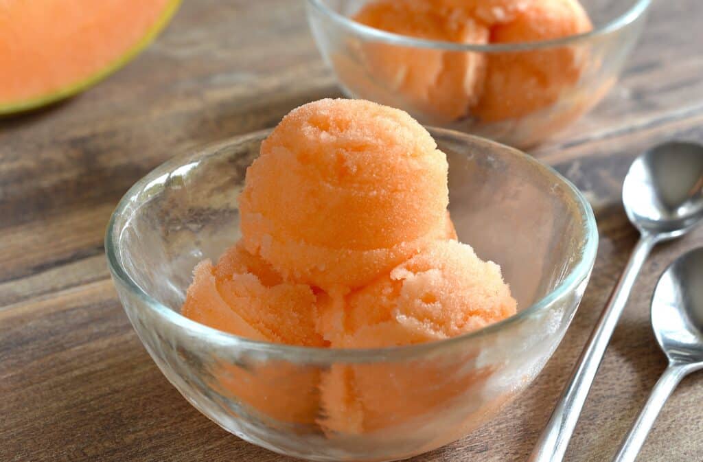 A close-up image of two bowls of melon sorbet on a wooden table with a piece of melon.