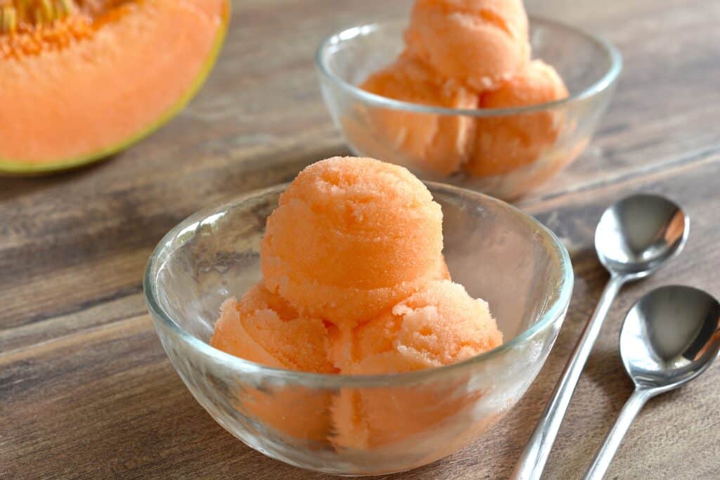 Two bowls of melon sorbet on a wooden table with a piece of melon and two spoons.