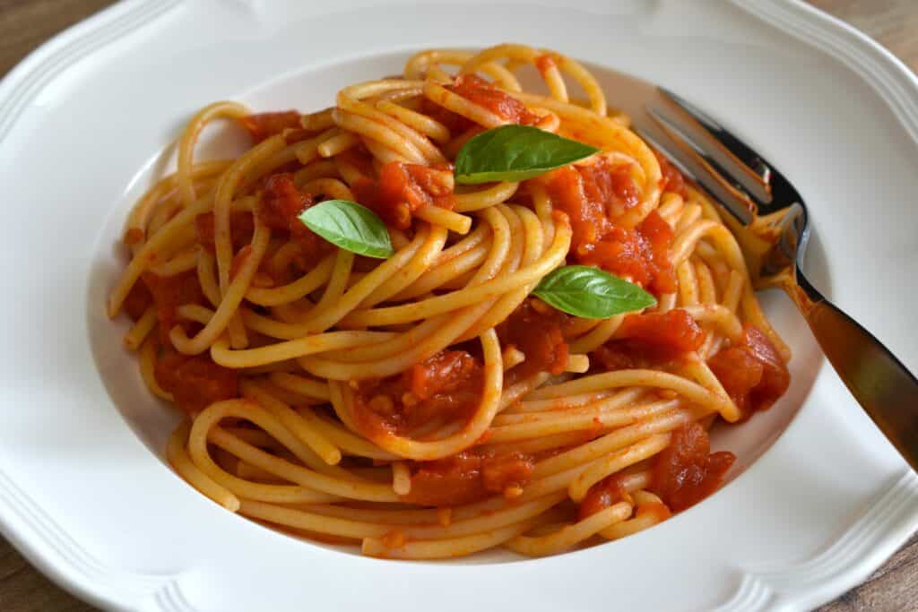 A close-up image of spaghetti with fresh tomato sauce on a white pasta plate with a for and basil leaf garnish.