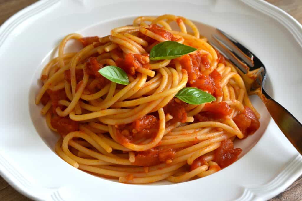 A close up image of spaghetti with fresh tomato sauce on a white pasta plate with a fork and basil leaf garnish.