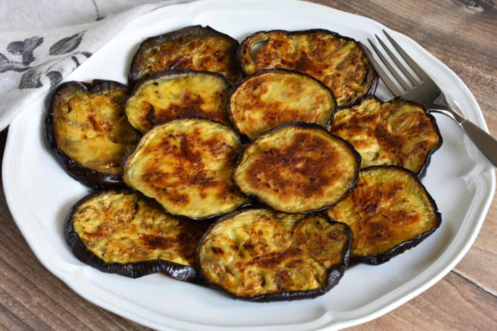 Roasted aubergine (eggplant) slices on a white plate with a fork and serviette.