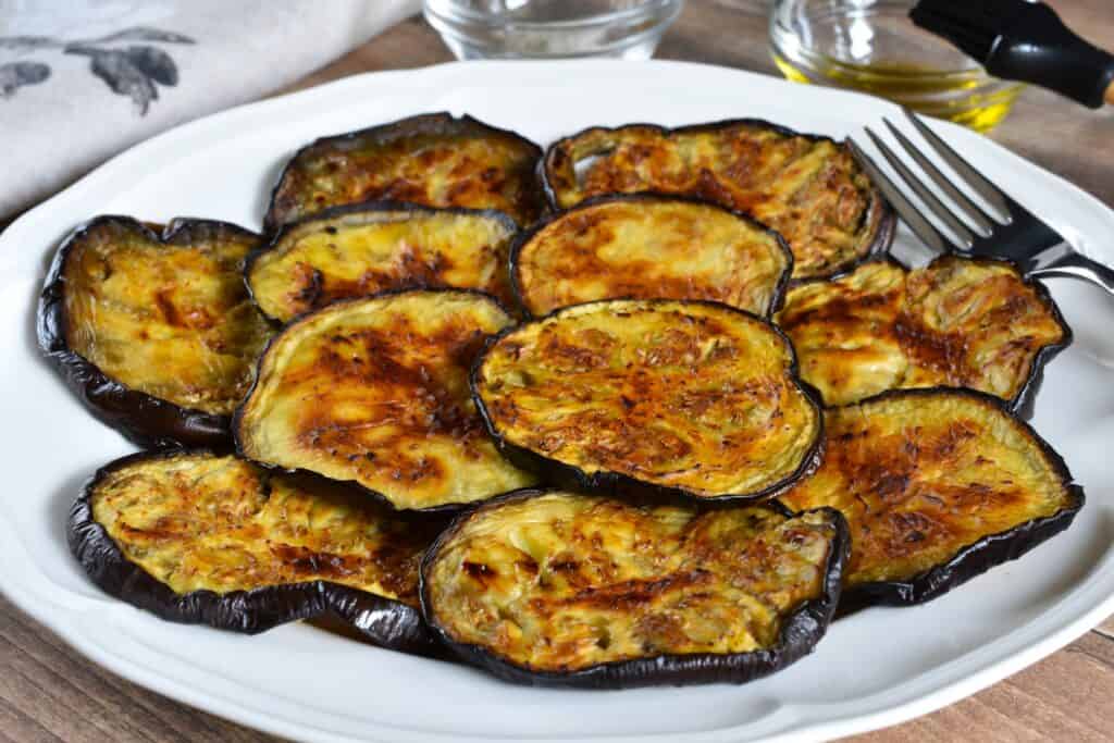 Roasted aubergine (eggplant) slices on a white plate with a fork and food props in the background.