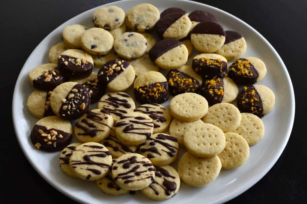 Vegan chocolate-dipped shortbread biscuits decorated with chocolate, hazelnuts, and orange zest, on a white plate.