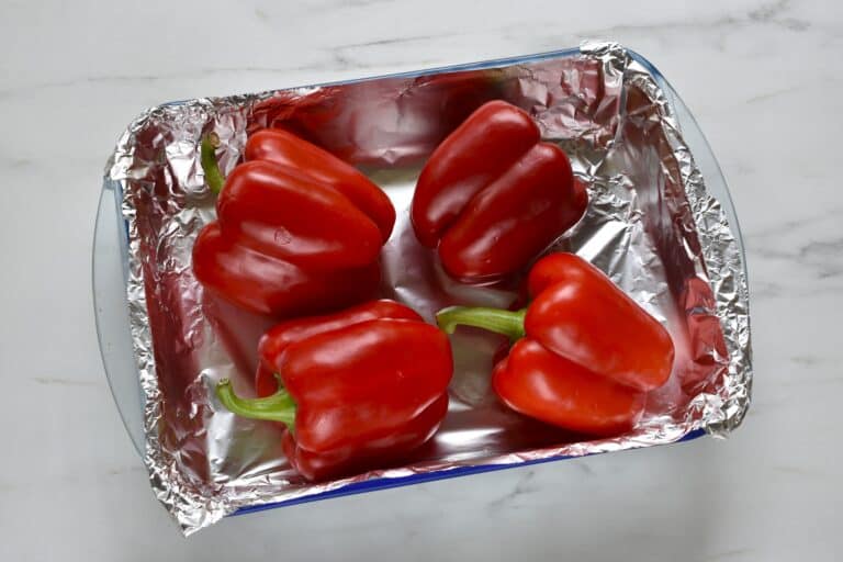 Four whole red peppers in an oven dish lined with aluminium foil.