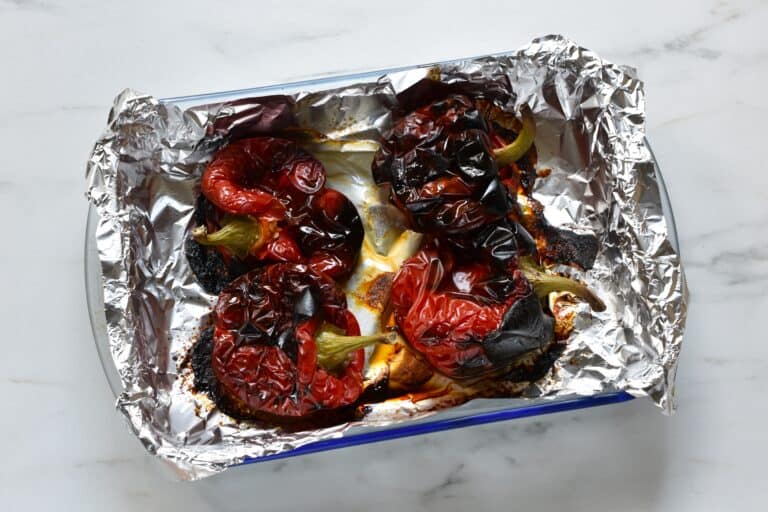 Four whole roasted red peppers in an oven dish lined with aluminium foil.