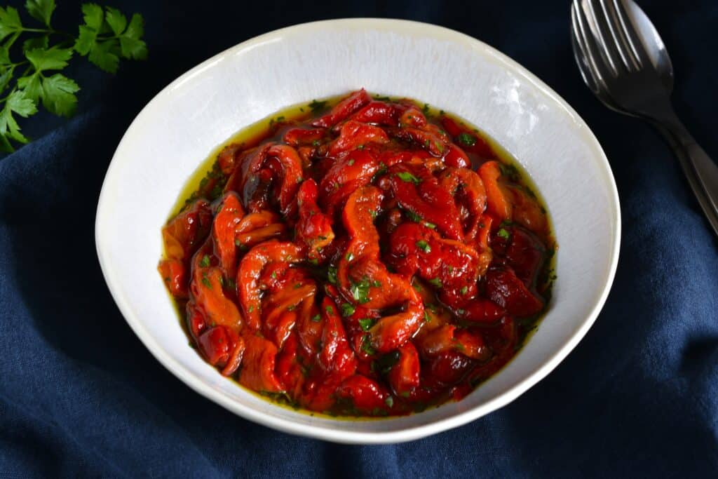 Roasted red pepper strips seasoned with extra virgin olive oil, salt, and chopped parsley, in a white bowl.